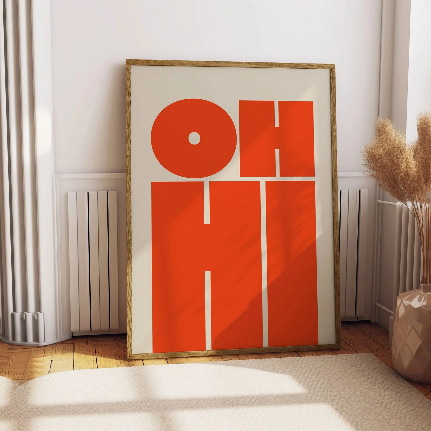 Affiche typographique orange vif avec les mots “OH HI”, posée au sol dans un intérieur lumineux, décoré de plantes séchées et parquet en bois clair.