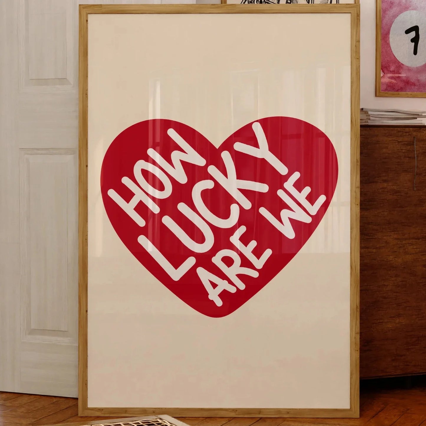 Affiche murale typographique avec message positif “How Lucky Are We” inscrit en blanc dans un cœur rouge, encadrée et posée contre un mur dans un intérieur chaleureux en bois, style cosy et vintage.
