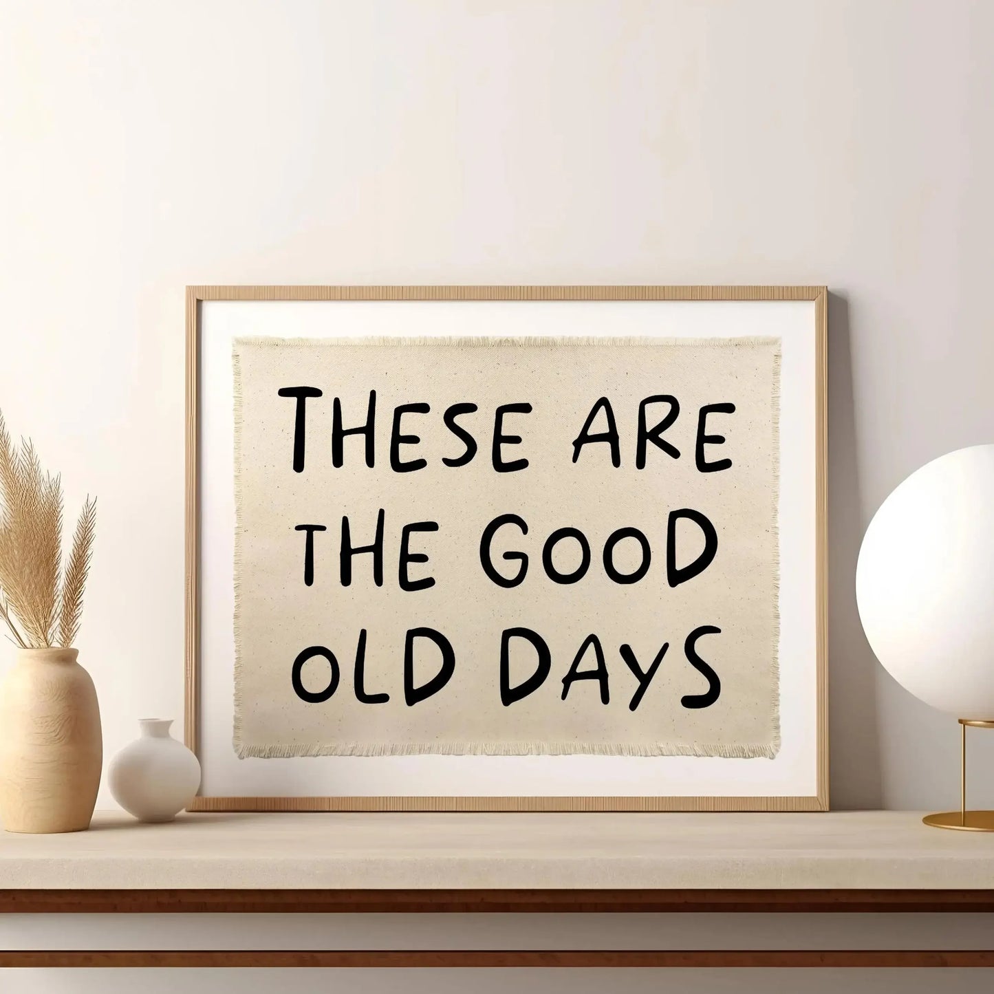 Affiche typographique “These Are The Good Old Days” au style manuscrit noir sur fond beige, encadrée et posée sur une console minimaliste avec objets déco naturels dans un intérieur lumineux et épuré.