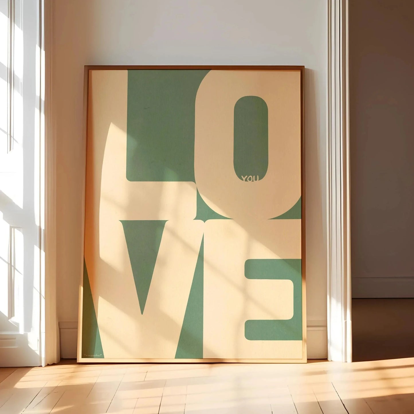 Affiche typographique design avec le mot “LOVE” en lettres majuscules géométriques vertes sur fond beige, encadrée en bois clair et exposée au sol dans une pièce baignée de lumière naturelle.