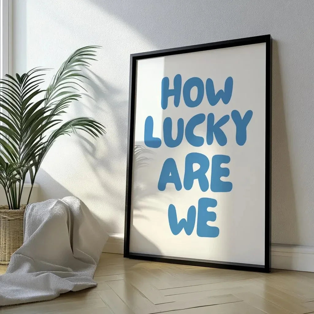 Affiche typographique moderne avec message “How Lucky Are We” en lettres bleues, encadrée en noir et posée contre un mur blanc, à côté d’une plante verte et d’un plaid dans un intérieur lumineux au style minimaliste