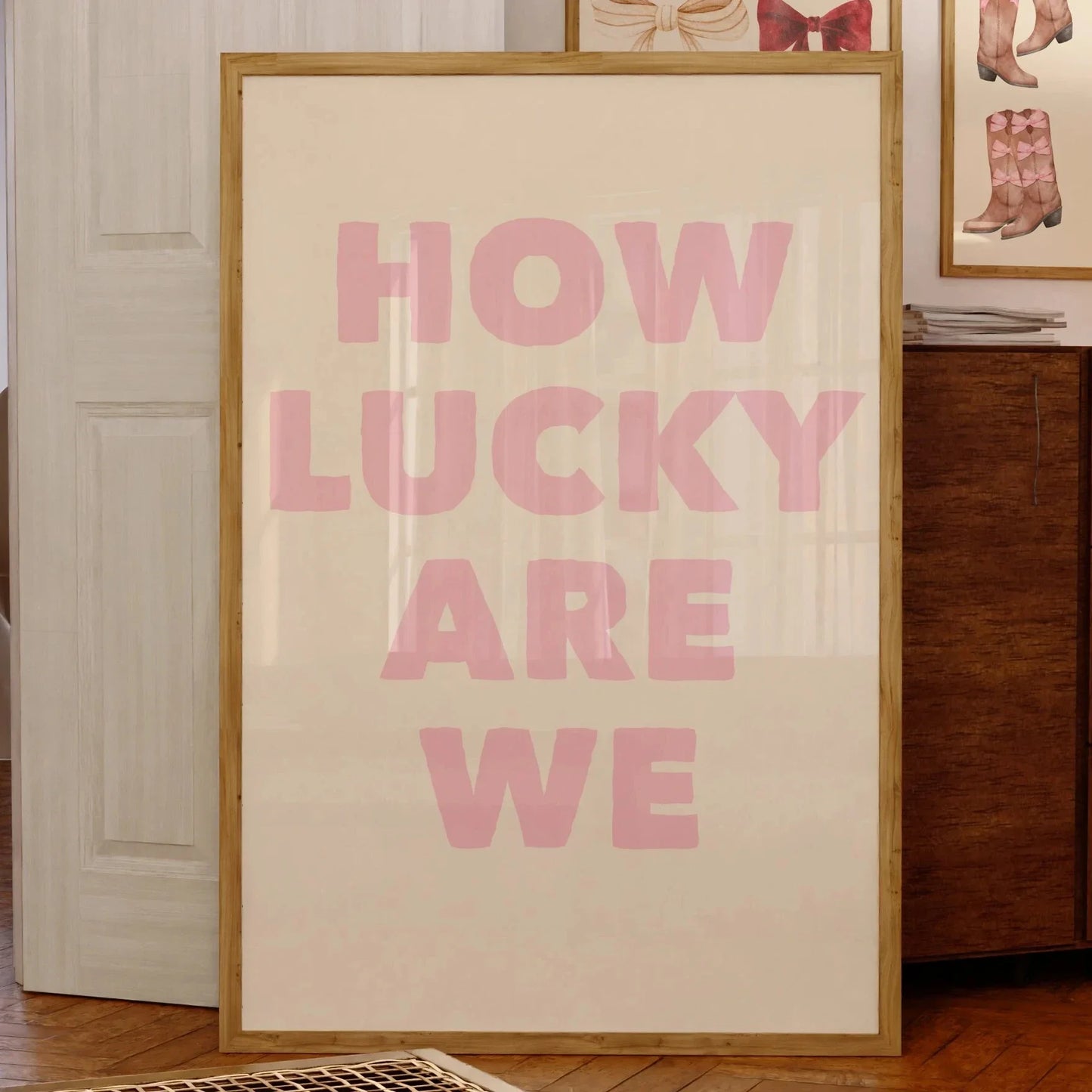 Affiche murale avec le message “HOW LUCKY ARE WE” en grandes lettres roses sur fond beige, encadrée bois clair, posée contre un mur dans un intérieur vintage avec illustrations encadrées en arrière-plan.