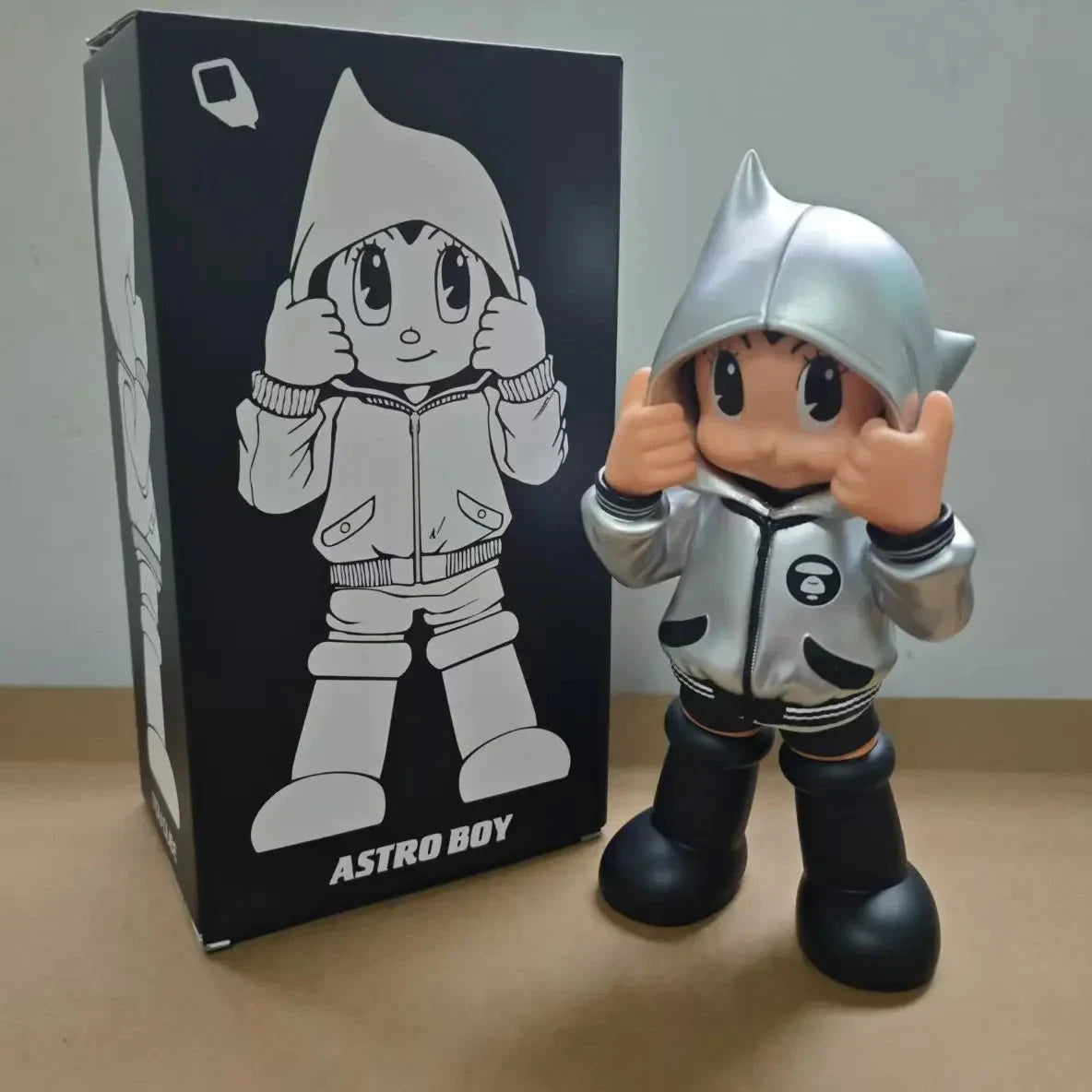 Figurine Astro Boy en hoodie argent pose mains levées, style street pop art avec boîte illustrée en arrière-plan