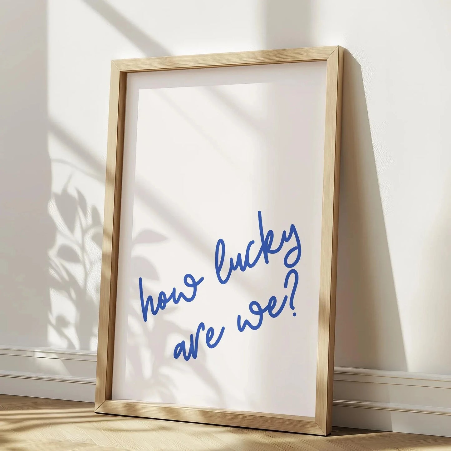 Affiche murale encadrée avec la citation manuscrite “how lucky are we?” en bleu, posée au sol dans un coin lumineux avec des ombres de plantes, déco minimaliste et moderne.