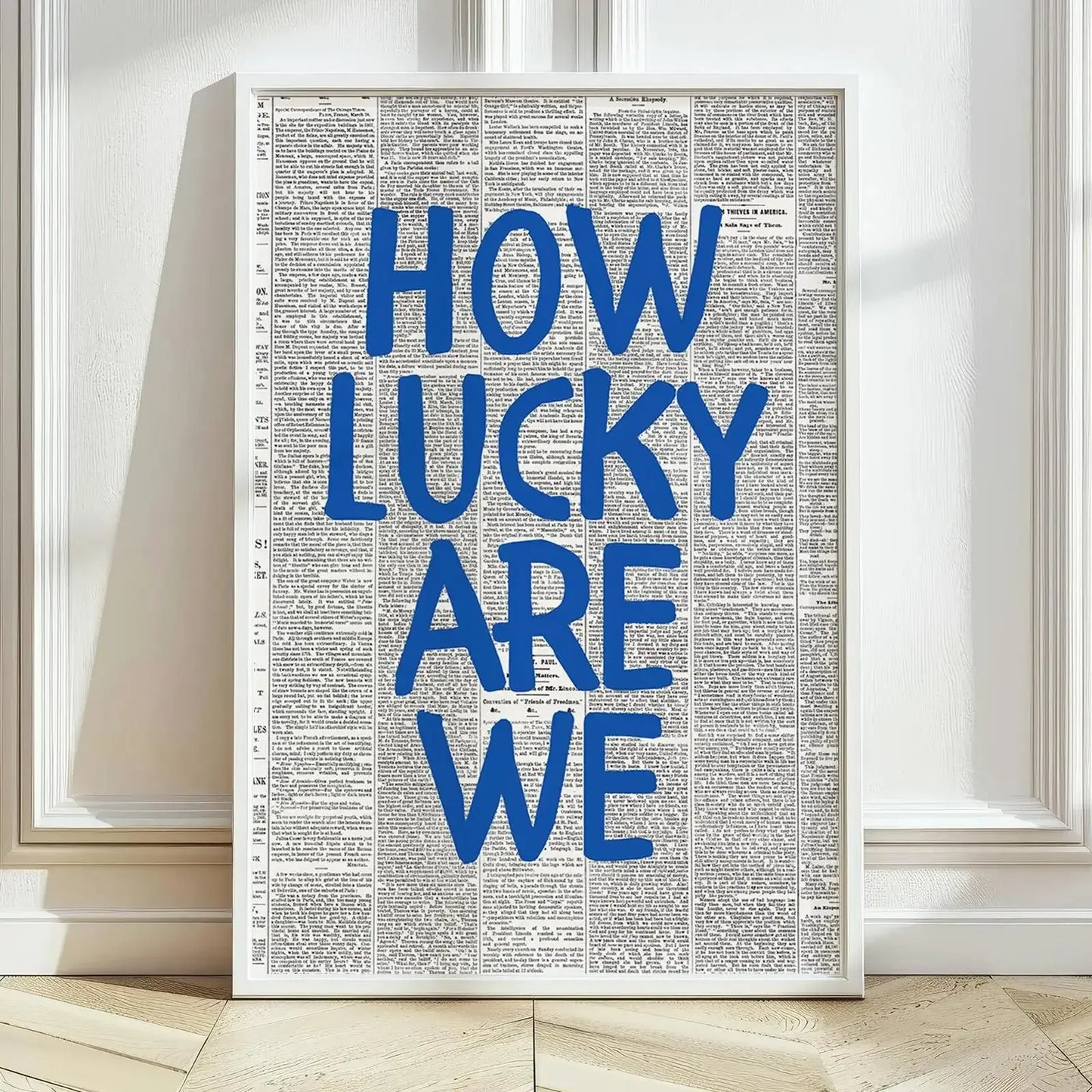 Affiche murale typographique “How Lucky Are We” en lettres bleues sur fond imitation journal, encadrée en blanc et posée au sol dans un intérieur lumineux au parquet clair.