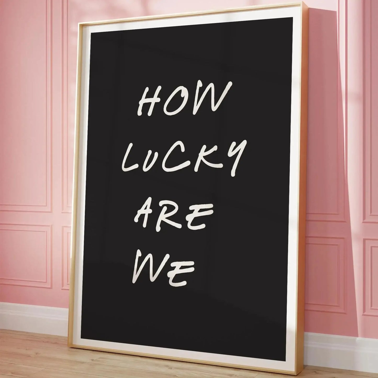Affiche décorative “How Lucky Are We” en lettres manuscrites blanches sur fond noir, encadrée en bois clair, posée au sol contre un mur rose à moulures dans un intérieur au style romantique et moderne.