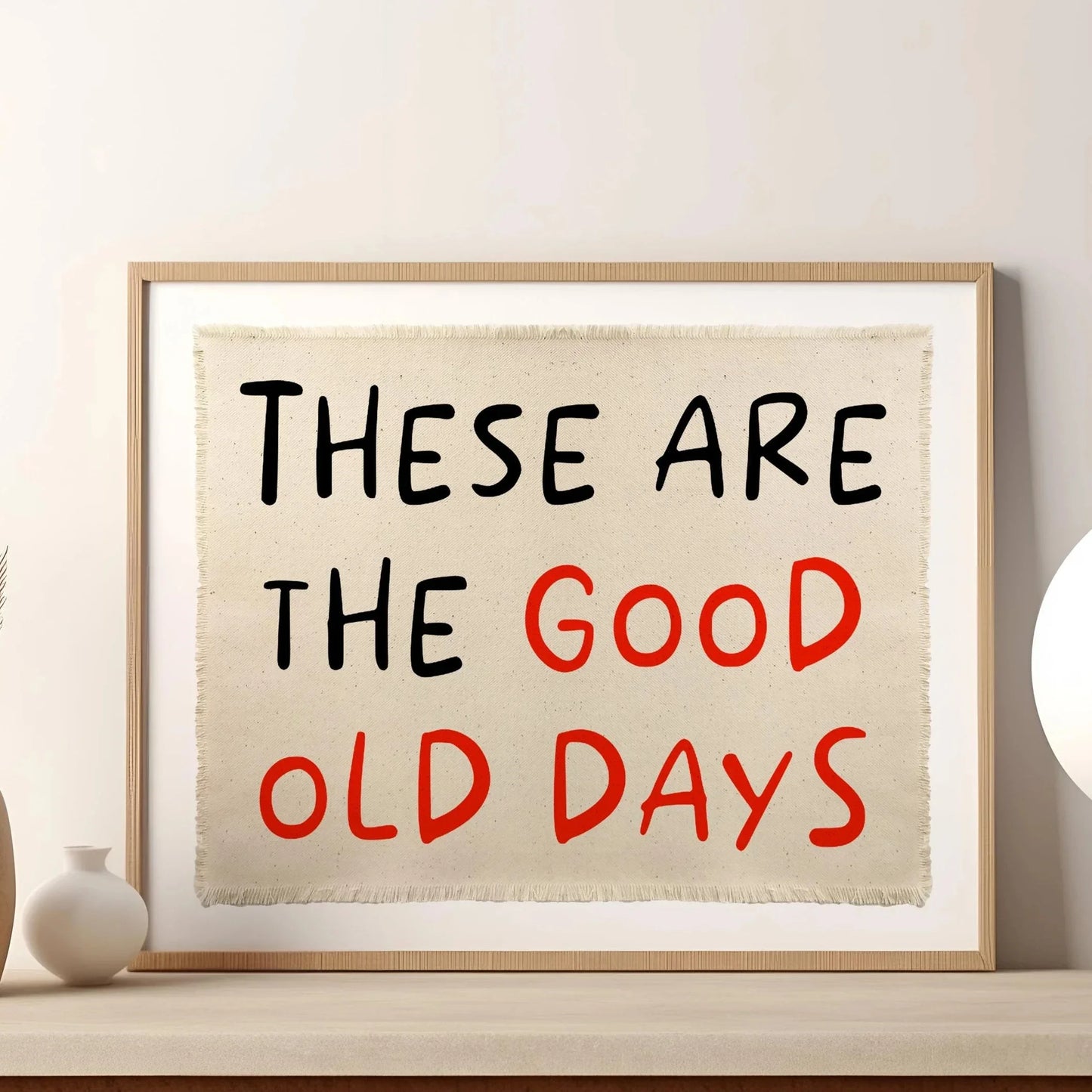 Affiche murale typographique “These Are The Good Old Days” avec lettrage noir et rouge sur fond toile beige, encadrée bois clair, posée sur une étagère avec vase déco dans un intérieur minimaliste et lumineux.