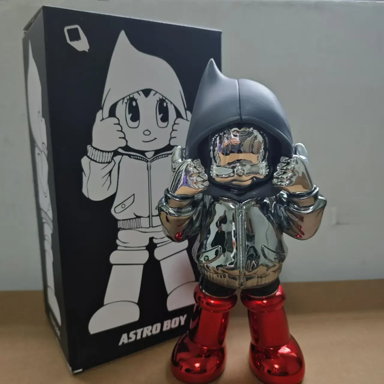 Figurine Astro Boy chromée en hoodie noir et bottes rouges, style chromé street art futuriste, présentée avec sa boîte illustrée en arrière-plan