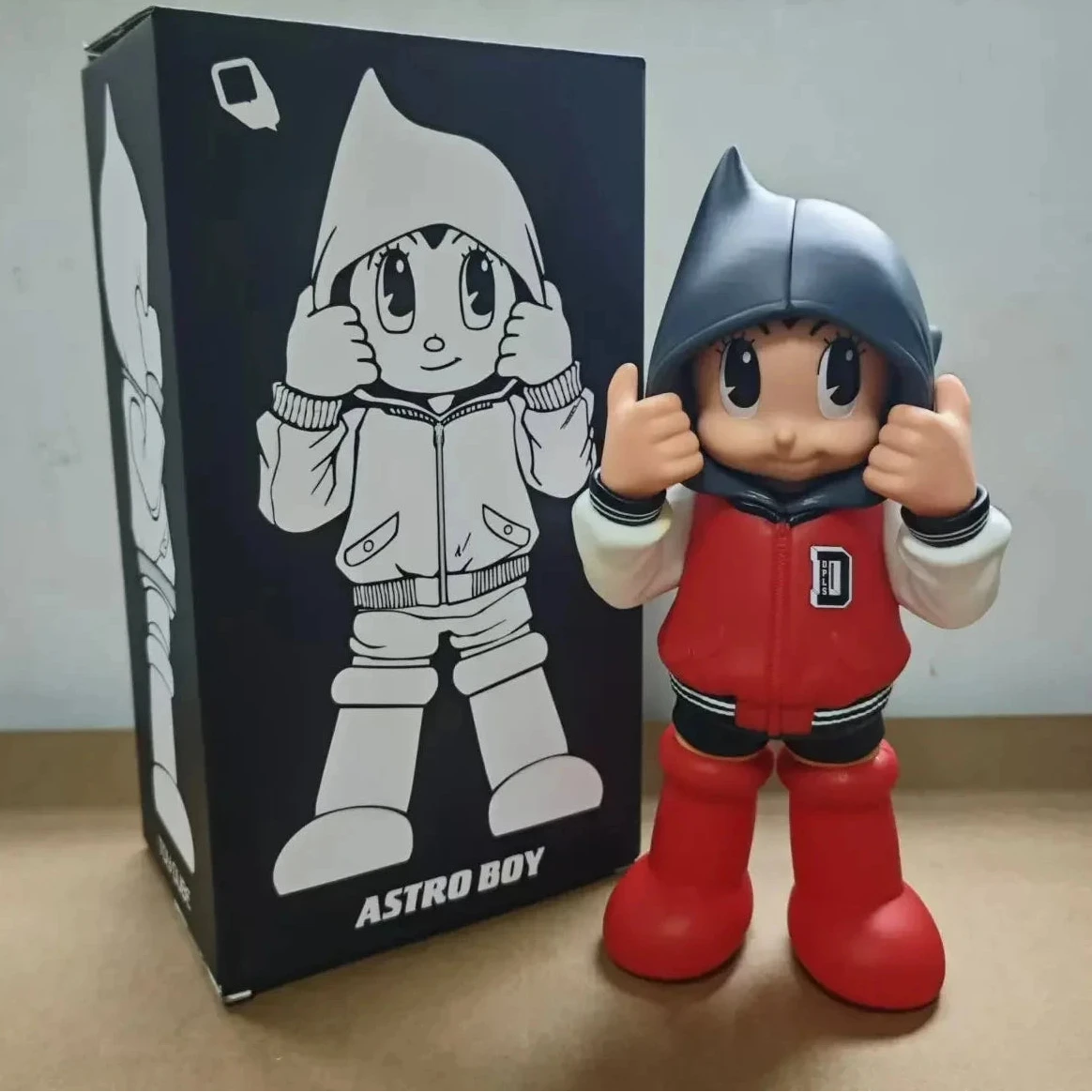 Figurine Astro Boy en hoodie noir et rouge, pose mains levées, style street pop art avec boîte illustrée en arrière-plan