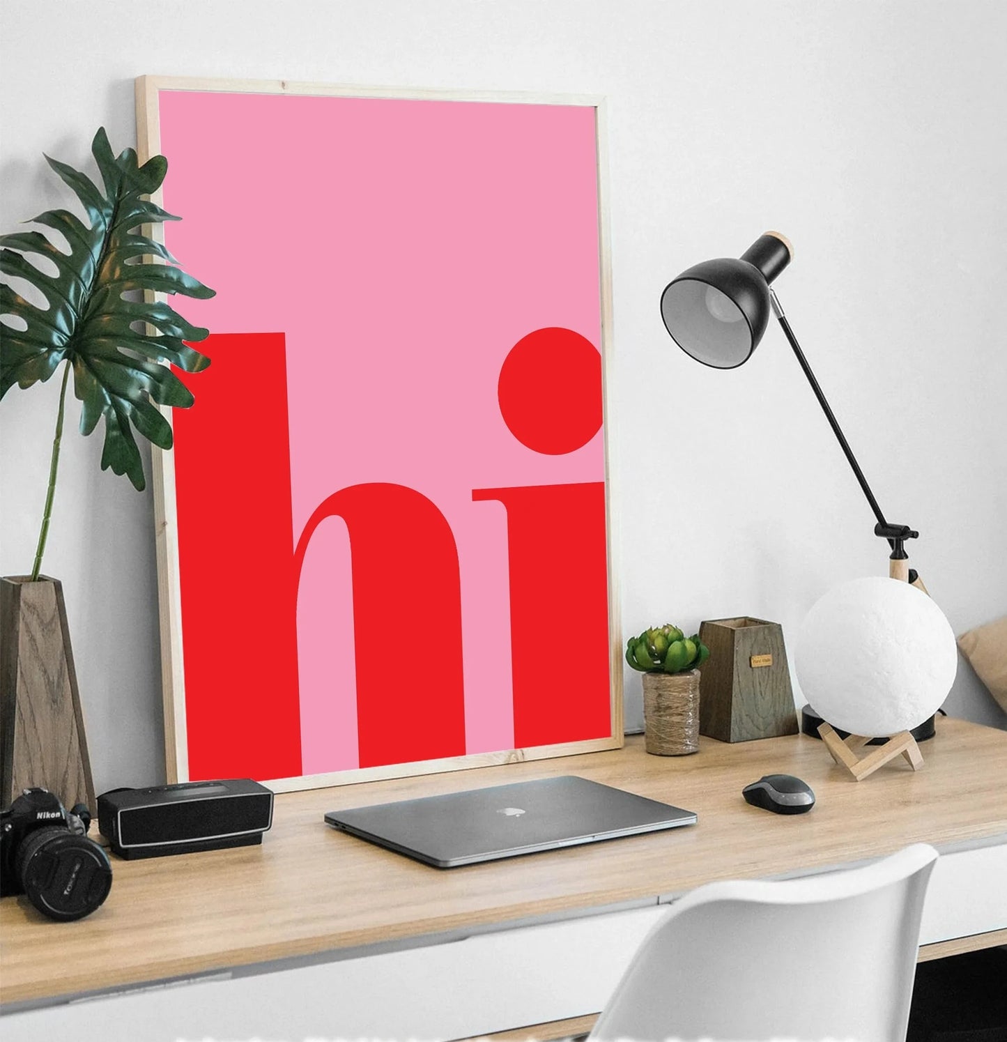 Affiche typographique rose et rouge avec le mot “hi”, posée sur un bureau moderne avec ordinateur portable, lampe design et plante verte, dans un intérieur lumineux et épuré.