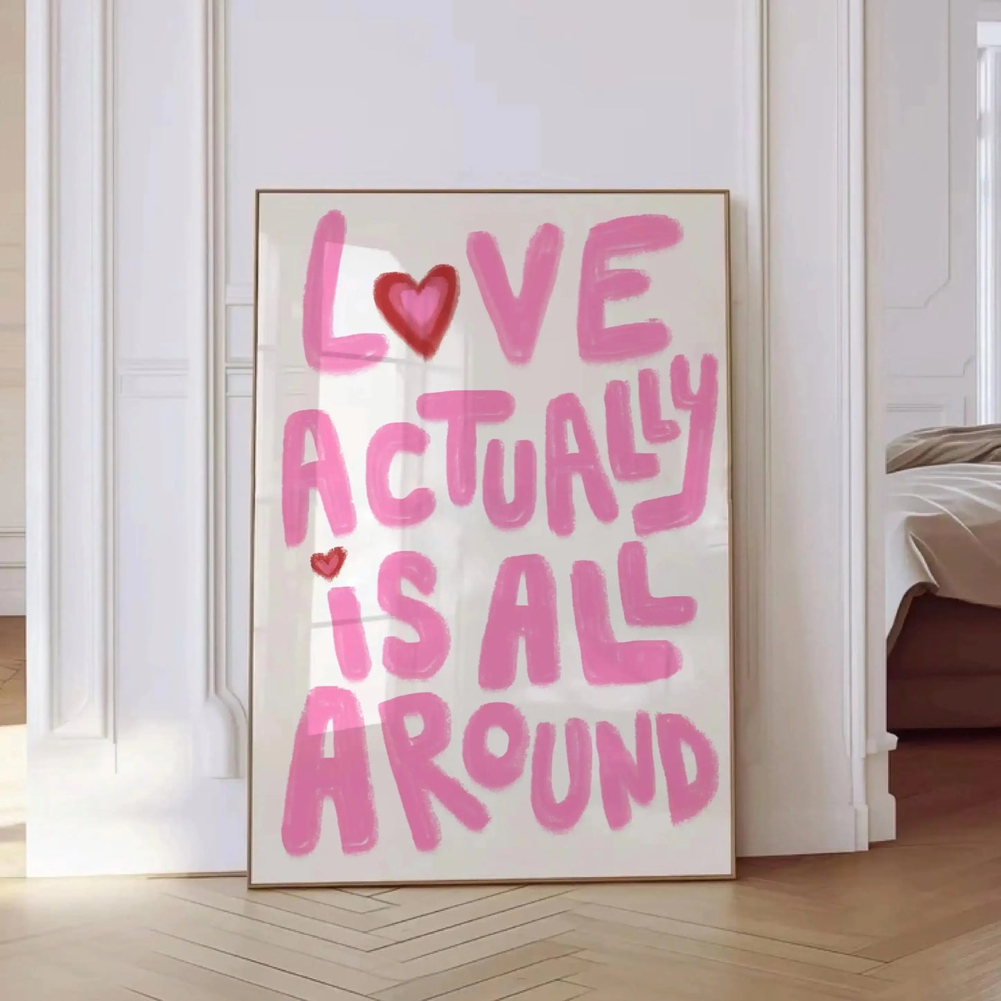 Affiche murale design avec le message “Love Actually Is All Around” écrit en lettres roses style manuscrit, encadrée en bois clair et exposée dans une pièce lumineuse au sol en parquet.