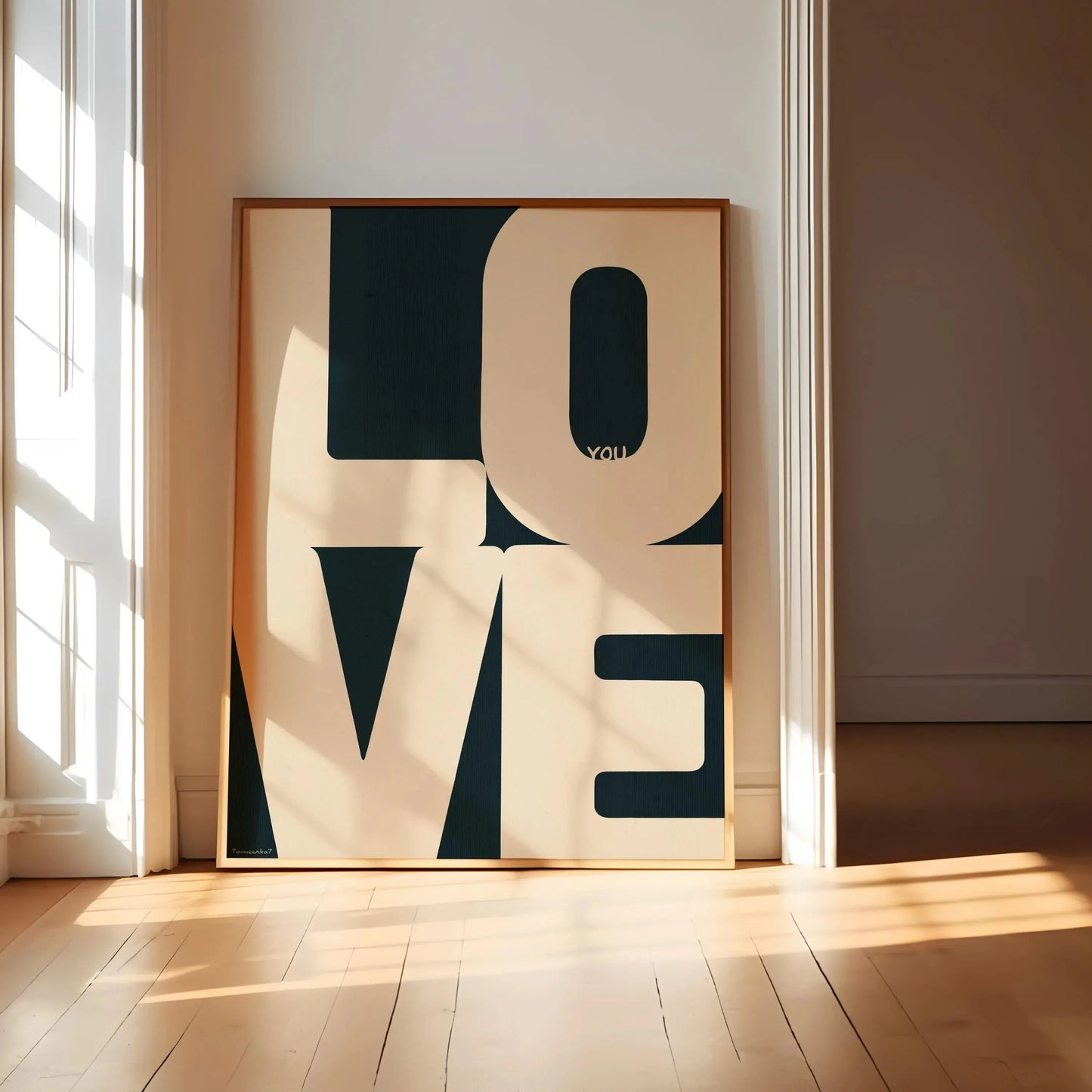 Affiche typographique moderne avec les lettres “LOVE” en majuscules crème sur fond noir, encadrée dans un cadre en bois naturel et posée dans une pièce lumineuse avec parquet bois clair et lumière du matin traversant une fenêtre à carreaux.
