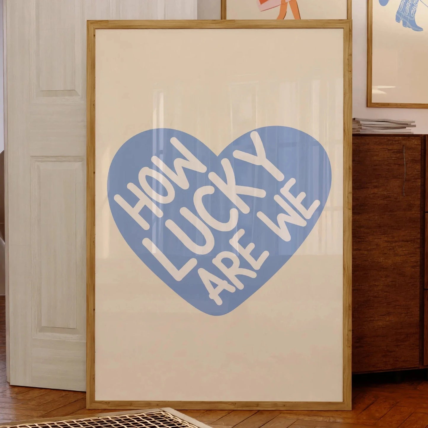 Affiche murale minimaliste avec le message “How Lucky Are We” en lettres blanches sur fond de cœur bleu, encadrée dans un intérieur au style vintage et chaleureux.