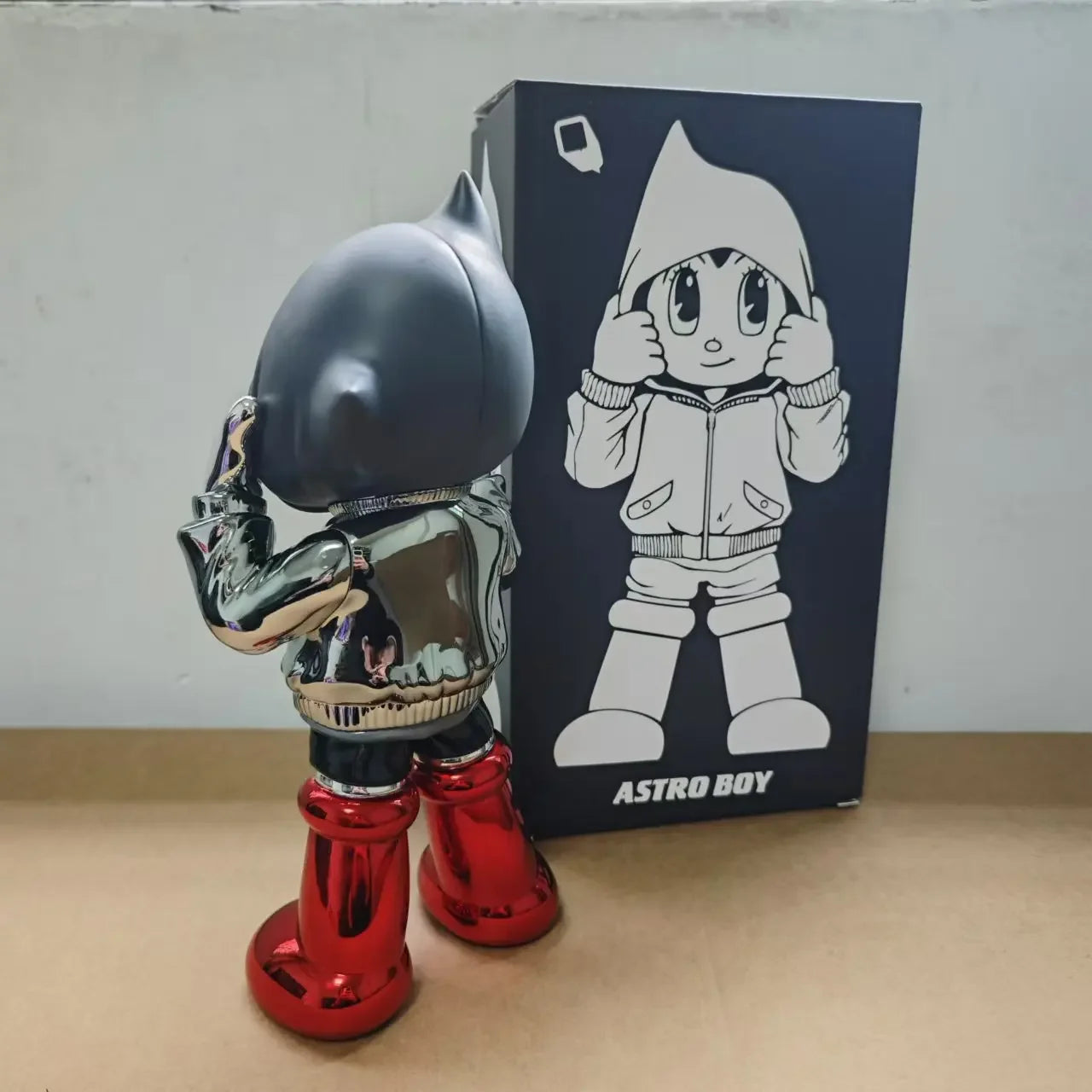 Figurine Astro Boy de dos , chromée en hoodie noir et bottes rouges, style street art futuriste, présentée avec sa boîte illustrée en arrière-plan