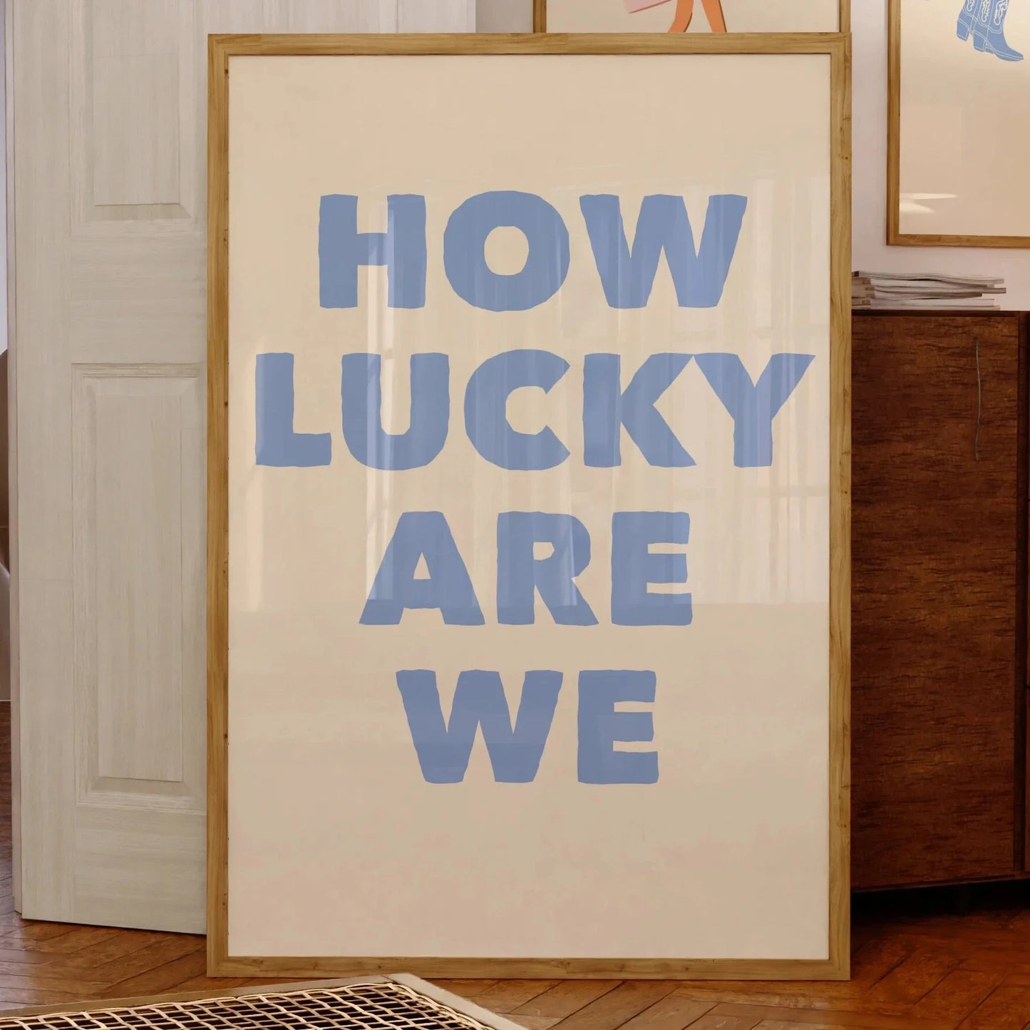 Affiche murale avec le message “HOW LUCKY ARE WE” en grandes lettres bleues sur fond beige, encadrée bois clair, posée contre un mur dans un intérieur vintage avec illustrations encadrées en arrière-plan.