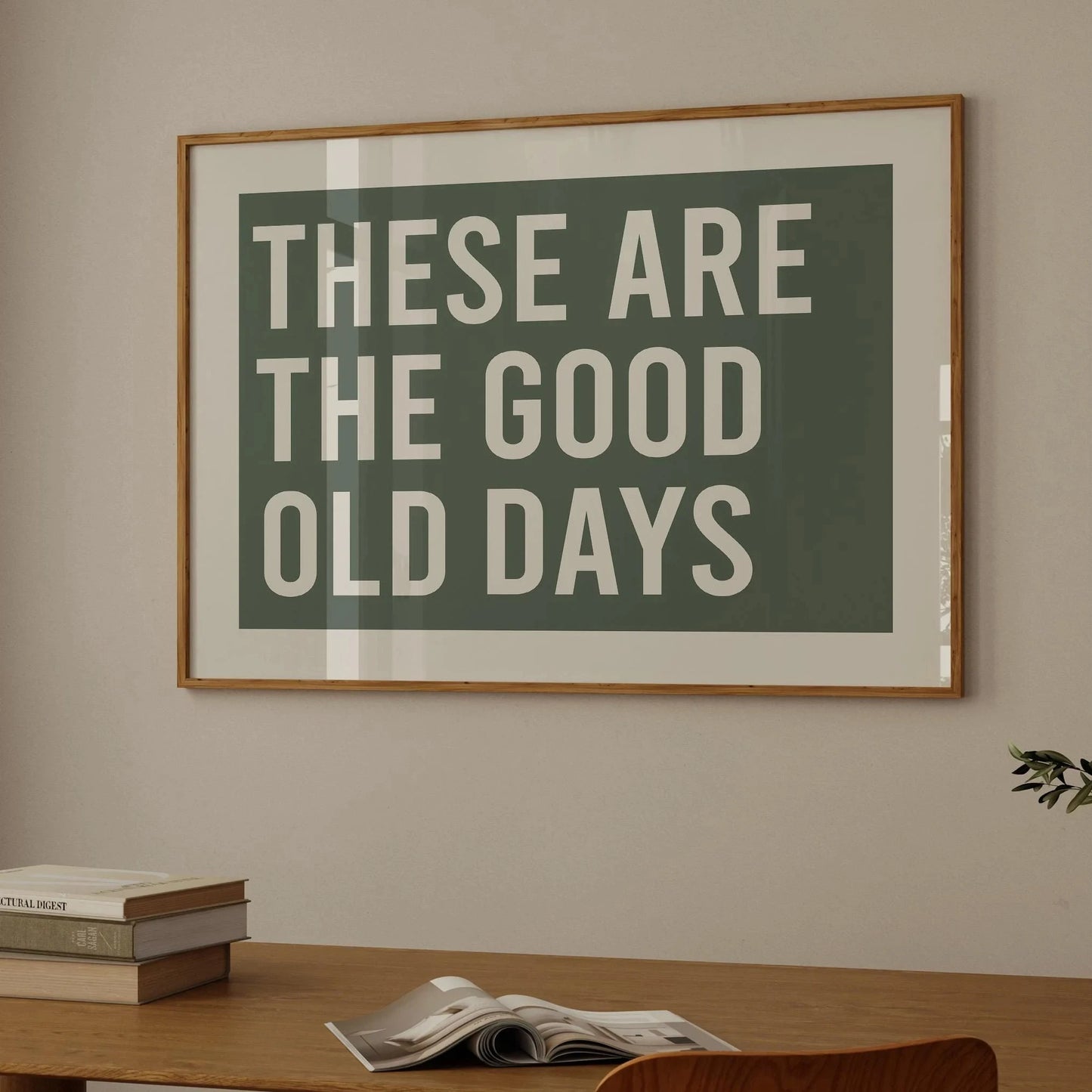 Affiche typographique “These Are The Good Old Days” en blanc sur fond vert kaki, encadrée bois clair, accrochée au mur au-dessus d’un bureau en bois dans un intérieur moderne et chaleureux.