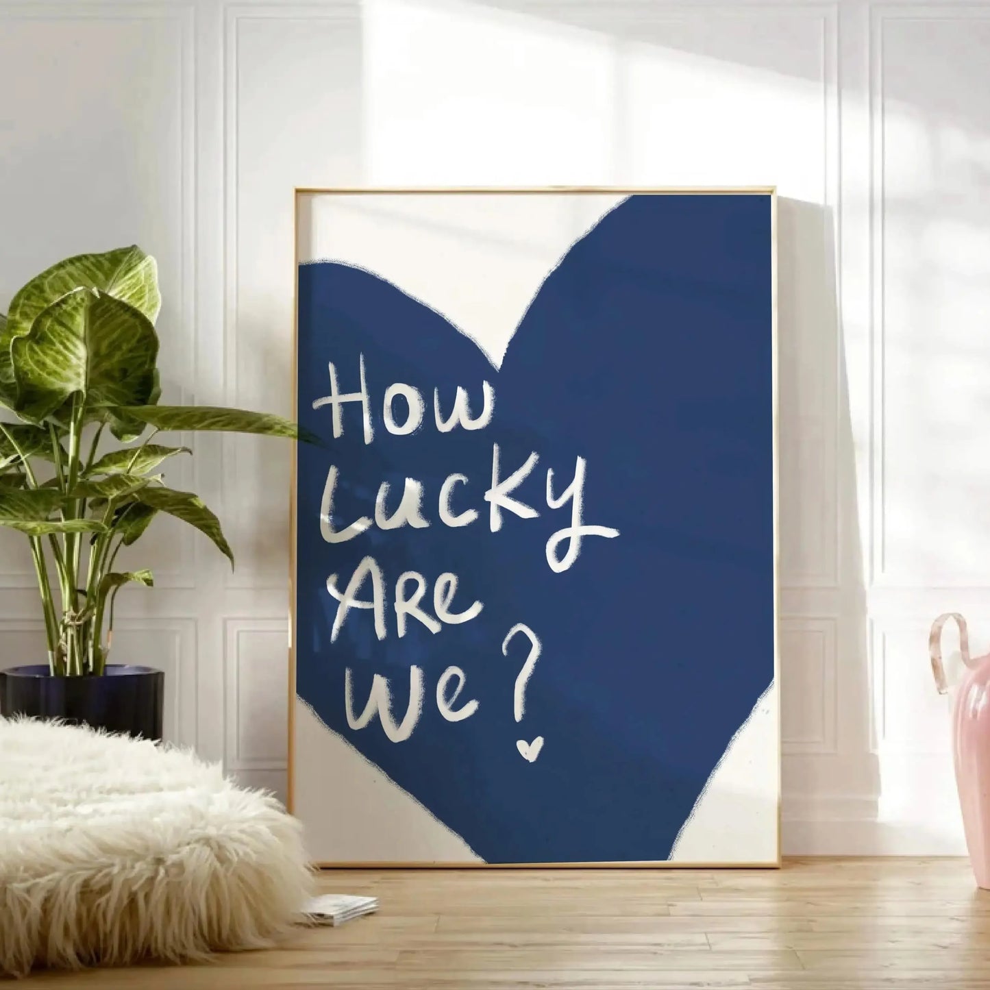 Affiche décorative encadrée avec un grand cœur bleu foncé et l’inscription manuscrite “How Lucky Are We?” en blanc, posée au sol dans un intérieur lumineux avec plante verte et déco cosy.