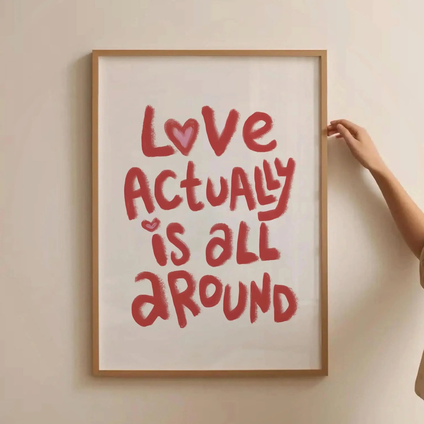 Affiche murale “Love Actually Is All Around” en lettres rouges manuscrites, avec petits cœurs, encadrée bois clair, tenue contre un mur beige par une main, dans un décor minimaliste.