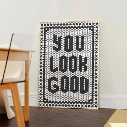 Affiche décorative typographique avec le message “YOU LOOK GOOD” formé par un motif de tuiles hexagonales noires et blanches, encadrée dans un cadre en bois clair, posée contre un mur blanc près d’une chaise design en bois et plexiglas.