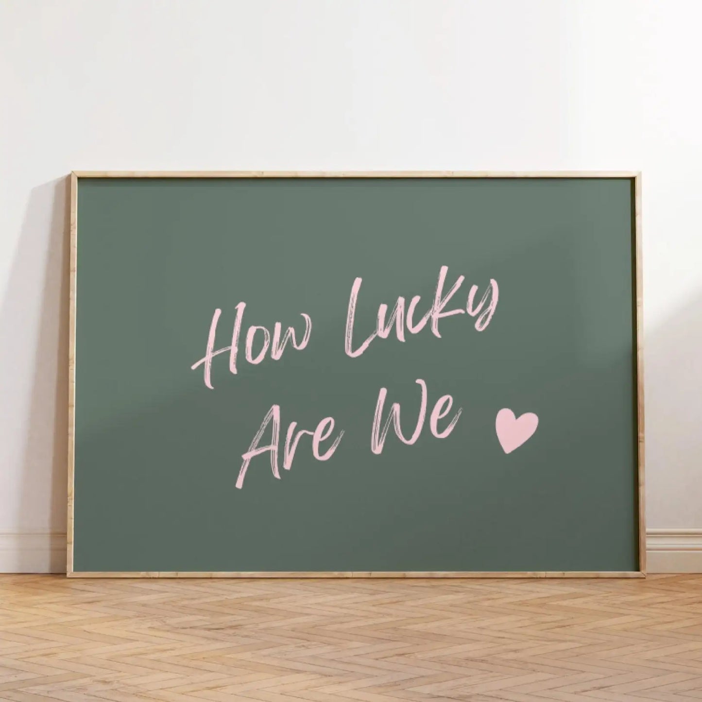 Affiche décorative au style moderne avec texte rose “How Lucky Are We” et cœur sur fond vert foncé, encadrée en bois clair et posée au sol dans un intérieur minimaliste et lumineux.