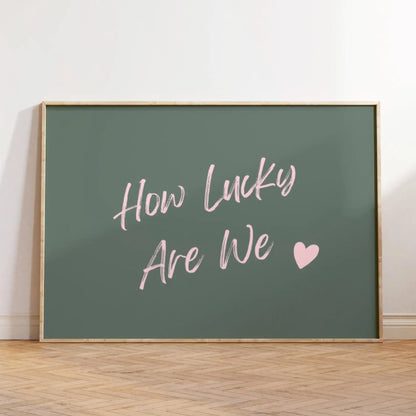 Affiche décorative au style moderne avec texte rose “How Lucky Are We” et cœur sur fond vert foncé, encadrée en bois clair et posée au sol dans un intérieur minimaliste et lumineux.