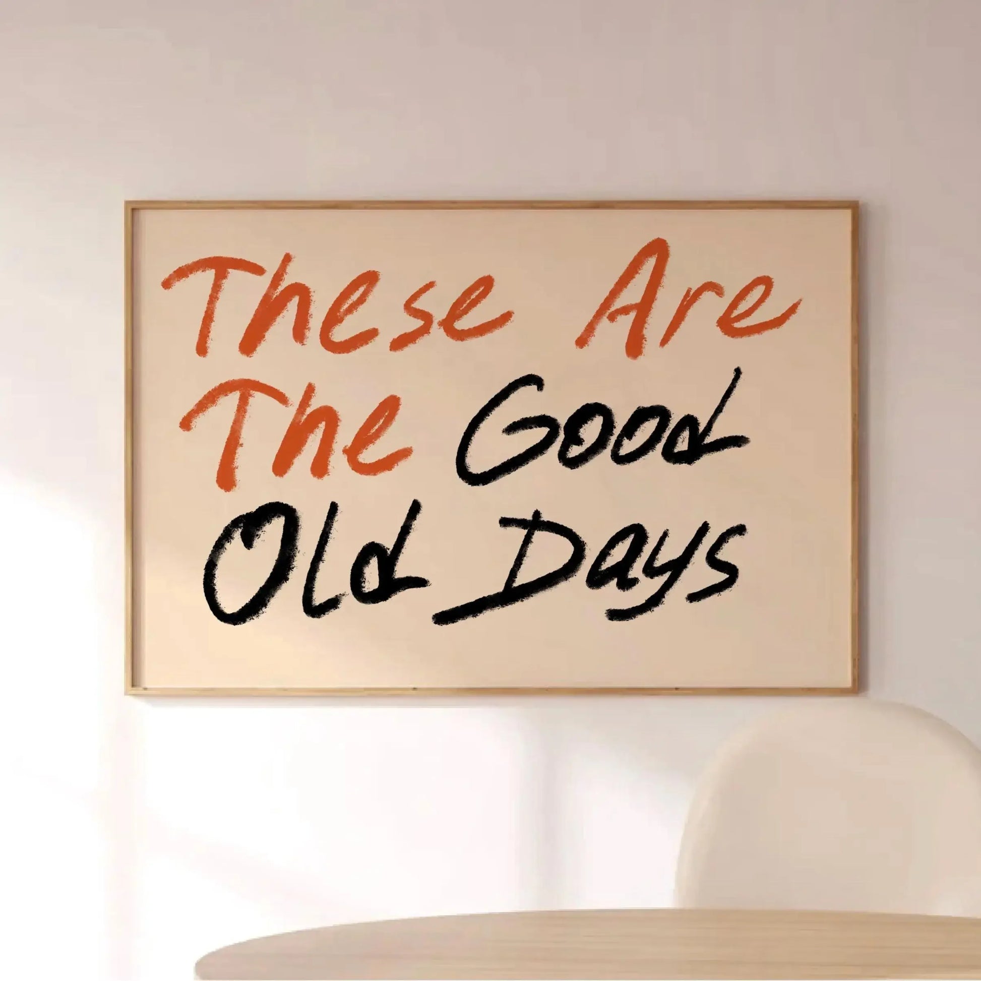 Affiche murale typographique avec le message “These Are The Good Old Days” en lettres manuscrites rouges et noires, encadrée et posée dans un intérieur moderne et lumineux avec chaise blanche.