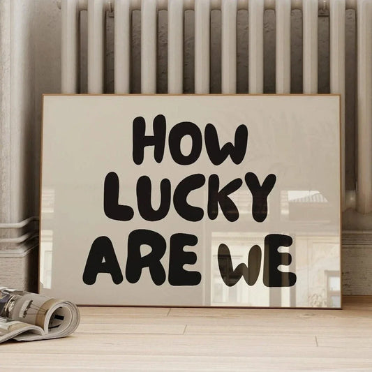 Affiche déco avec le message “How Lucky Are We” en typographie noire épaisse, posée au sol devant un radiateur rétro dans un intérieur au style brut et contemporain.