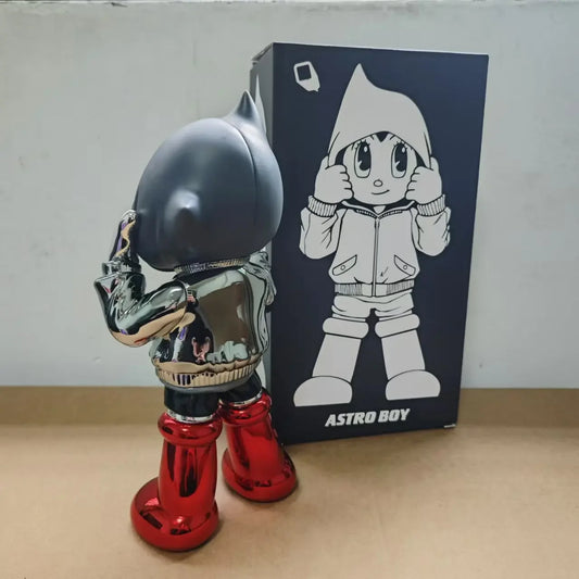 Figurine Astro Boy de dos , chromée en hoodie noir et bottes rouges, style street art futuriste, présentée avec sa boîte illustrée en arrière-plan