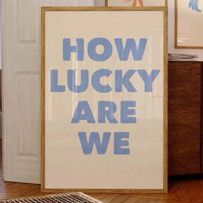 Affiche murale avec le message “HOW LUCKY ARE WE” en grandes lettres bleues sur fond beige, encadrée bois clair, posée contre un mur dans un intérieur vintage avec illustrations encadrées en arrière-plan.