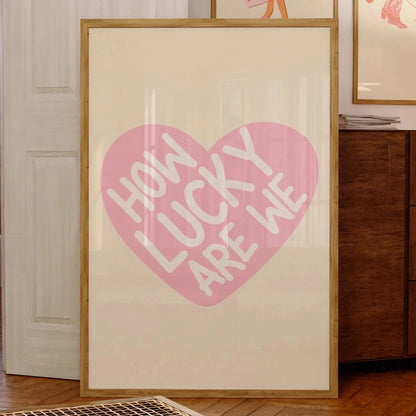 Affiche murale typographique avec message positif “How Lucky Are We” inscrit en blanc dans un cœur rose, encadrée et posée contre un mur dans un intérieur chaleureux en bois, style cosy et vintage.