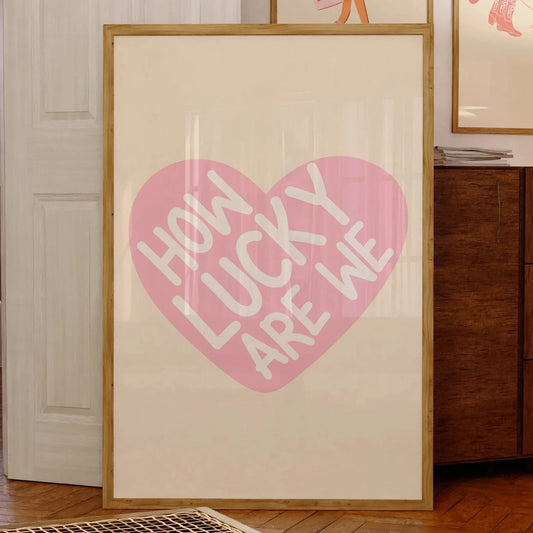 Affiche murale typographique avec message positif “How Lucky Are We” inscrit en blanc dans un cœur rose, encadrée et posée contre un mur dans un intérieur chaleureux en bois, style cosy et vintage.